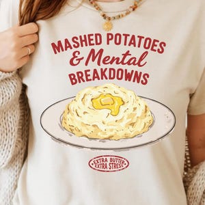 Puede incluir: Camiseta blanca con el texto "Mashed Potatoes & Mental Breakdowns" en rojo. Debajo del texto hay una imagen de dibujos animados de puré de patatas con una porción de mantequilla en la parte superior. Un círculo rojo con el texto "Extra Butter Extra Stress" está en la parte inferior de la camiseta.