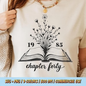 May include: A white t-shirt with a black and white design of a book with flowers growing out of it. The text "chapter forty" is written below the book. The numbers "19" and "85" are on either side of the book.