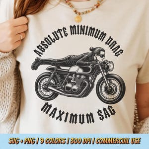May include: A cream-colored t-shirt featuring a black graphic of a person lying flat on a motorcycle. The text "ABSOLUTE MINIMUM DRAG" is arched above the illustration, and "MAXIMUM SAG" is printed in a gothic font below the motorcycle.