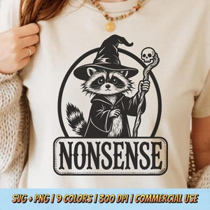 May include: A cream-colored t-shirt featuring a black and white graphic of a raccoon dressed as a wizard, holding a staff with a skull. The word "NONSENSE" is displayed below the raccoon in a rectangular frame.