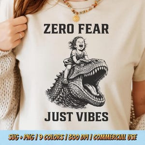 Op de afbeelding: Beige T-shirt met de tekst "ZERO FEAR JUST VIBES" boven een zwart-wit illustratie van een kind dat op een dinosaurus rijdt. Het ontwerp bevat SVG- en PNG-formaten, 9 kleuren, 300 DPI en commercieel gebruik.