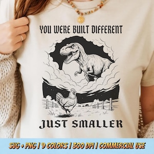 May include: Cream-coloured t-shirt featuring a black and white illustration of a T-Rex and a chicken. The text above reads "YOU WERE BUILT DIFFERENT", and below, "JUST SMALLER". The bottom of the shirt has text that reads "SVG + PNG | 9 COLORS | 300 DPI | COMMERCIAL USE".