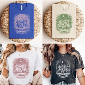 May include: Four t-shirts in blue, beige, white, and charcoal gray, each featuring a graphic of dancing figures under a crescent moon and the text "GOD FORBID WOMEN HAVE HOBBIES". The shirts are folded on woven mats.