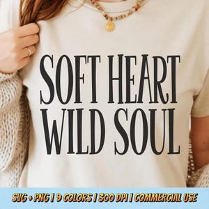 May include: A cream-colored t-shirt with the words "SOFT HEART WILD SOUL" in bold black letters. The shirt is being worn by a person. A necklace with a sun pendant is visible. The bottom of the image includes text: SVG + PNG | 9 COLORS | 300 DPI | COMMERCIAL USE.