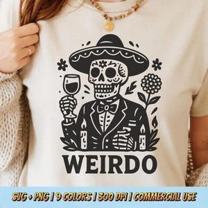 May include: Beige t-shirt featuring a black graphic of a skeleton wearing a sombrero and tuxedo, holding a wine glass. The word "WEIRDO" is printed in bold letters below the graphic. The design includes floral and candle elements.