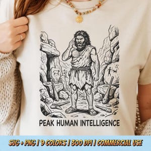 May include: Beige t-shirt featuring a black and white illustration of a caveman holding a phone to his ear, with the text "PEAK HUMAN INTELLIGENCE" below. The design includes cave paintings and stone tools. The bottom of the shirt has text that reads "SVG + PNG | 9 COLORS | 300 DPI | COMMERCIAL USE".