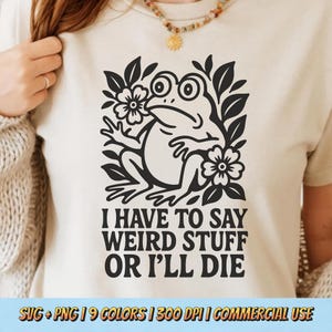 May include: Beige t-shirt featuring a black graphic of a frog surrounded by flowers and leaves. The text "I HAVE TO SAY WEIRD STUFF OR I'LL DIE" is printed below the frog. The shirt is a neutral color.