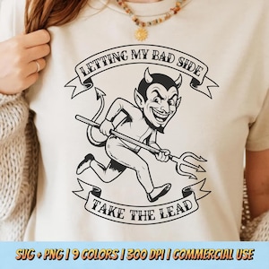 May include: A cream-colored t-shirt features a black and white cartoon devil running with a pitchfork. The text on the shirt reads "LETTING MY BAD SIDE TAKE THE LEAD" in a banner design. The shirt is a casual wear item.
