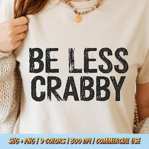 May include: A cream-colored t-shirt with the phrase "BE LESS CRABBY" printed in large, distressed black letters. The shirt is a casual style, suitable for everyday wear. The bottom of the image includes text about the design's availability.