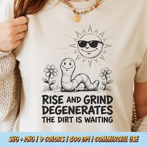 Pode incluir: Uma t-shirt cor de creme com um gráfico de linhas pretas de um verme sorridente e um sol com óculos de sol. O texto diz: RISE AND GRIND DEGENERATES THE DIRT IS WAITING. O design inclui duas pequenas flores ao lado do verme.