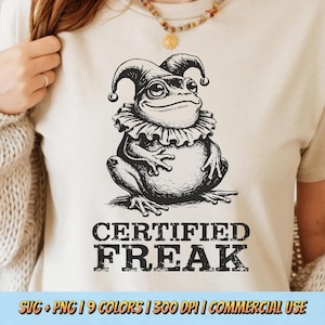 May include: Cream-colored t-shirt featuring a black and white illustration of a frog wearing a jester's hat and collar. The text "CERTIFIED FREAK" is printed below the frog. The shirt is a casual wear item.