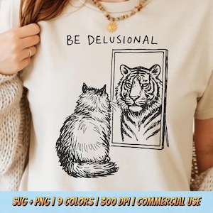 May include: Cream-colored t-shirt with the text "BE DELUSIONAL" above a drawing of a cat looking in a mirror at a tiger. The cat is drawn in black and white, as is the tiger in the mirror. The shirt is being worn.