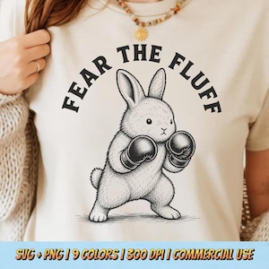 Op de afbeelding: Een crèmekleurig t-shirt met een zwarte grafische illustratie van een konijn met bokshandschoenen aan. De tekst "FEAR THE FLUFF" is in een vet, gebogen zwart lettertype boven het konijn op de borst van het shirt gedrukt.