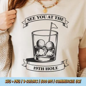 Może przedstawiać: Kremowa koszulka z czarną grafiką szklanki do koktajli z piłeczkami golfowymi i kostkami lodu. Tekst "SEE YOU AT THE 19TH HOLE" jest wyświetlany nad i pod szklanką w formie banera. Flaga golfowa znajduje się w szklance.