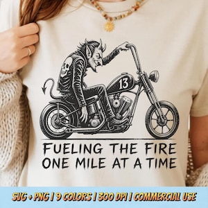 May include: A cream-colored t-shirt featuring a black graphic of a horned devil riding a motorcycle with the number 13 on the gas tank. Below the motorcycle, the text reads: FUELING THE FIRE ONE MILE AT A TIME. The design is printed in a dark ink.