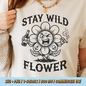 May include: A cream-colored t-shirt features a black graphic of a winking flower holding a beer bottle and a lit cigar. The text "STAY WILD FLOWER" is above and below the flower. The shirt has a crew neck and short sleeves.