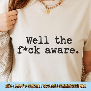 Pode incluir: Camiseta creme com a frase "Well the f*ck aware" impressa em preto, em negrito. A camiseta faz parte de uma coleção de roupas e é adequada para uso casual. A imagem também inclui texto na parte inferior.