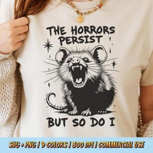 以下が含まれることがあります： 「The Horrors Persist But So Do I」というテキストと、オポッサムの白黒イラストが描かれた白いTシャツです。