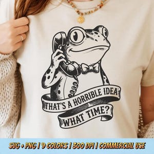 Könnte beinhalten: Ein cremefarbenes T-Shirt mit einer Schwarz-Weiß-Illustration eines Frosches mit Fliege und Telefon. Der Text auf dem Shirt lautet: "THAT'S A HORRIBLE IDEA WHAT TIME?"