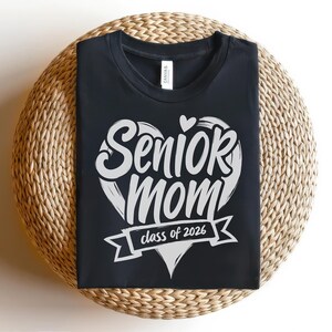 May include: Black t-shirt with a white heart-shaped design that reads "Senior Mom" with a small heart above the word "Senior". Below the heart is a banner that says "class of 2026". The shirt is displayed on a woven, circular surface.
