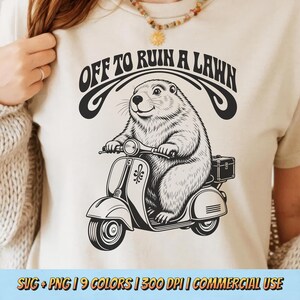 May include: A cream-colored t-shirt featuring a black graphic of a groundhog riding a scooter. Above the groundhog, the text reads OFF TO RUIN A LAWN in a bold, arched font. The design includes a small storage box on the back of the scooter.