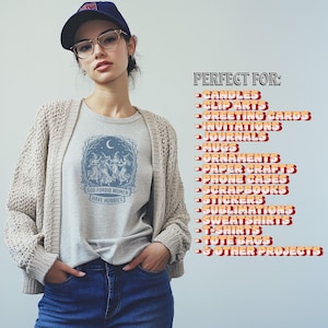 May include: A light gray t-shirt with the text "GOD FORBID WOMEN HAVE HOBBIES" and a graphic of dancing figures under a crescent moon. The image also shows a woman wearing a beige cardigan, blue jeans, and a navy blue cap.