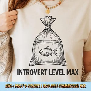 May include: Beige t-shirt featuring a black and white illustration of a fish in a bag of water. The text "INTROVERT LEVEL MAX" is printed below the graphic. The shirt is a casual style.