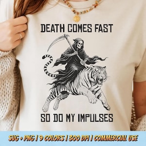 May include: A cream-colored t-shirt featuring a black graphic of a skeleton in a hooded robe holding a scythe while riding a tiger. The text above reads DEATH COMES FAST and the text below reads SO DO MY IMPULSES in a bold, dark font.