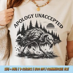 以下が含まれることがあります： ベージュのTシャツ。黒と白のフラミンゴのグラフィックと「APOLOGY UNACCEPTED」の文字。デザインには森、湖、太陽が含まれています。シャツの下部には「SVG + PNG | 9 COLORS | 300 DPI | COMMERCIAL USE」と書かれています。