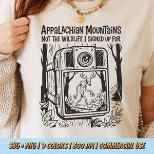 May include: Cream-coloured t-shirt featuring a black graphic of a trail camera with a skeletal figure. Text reads "APPALACHIAN MOUNTAINS NOT THE WILDLIFE I SIGNED UP FOR". The shirt has a crew neck and short sleeves.