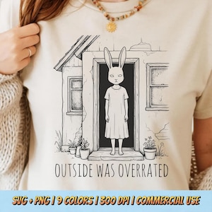 Könnte beinhalten: Beigefarbenes T-Shirt mit einer Schwarz-Weiß-Illustration einer kaninchenartigen Figur, die in einer Tür steht. Der Text "OUTSIDE WAS OVERRATED" ist darunter gedruckt. Das Design umfasst ein kleines Haus und Topfpflanzen.