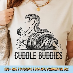 May include: A beige t-shirt featuring a black graphic illustration of a person wrestling a large snake while holding a knife. Below the illustration, the text CUDDLE BUDDIES is printed in a bold, dark font. The design is centred on the chest.