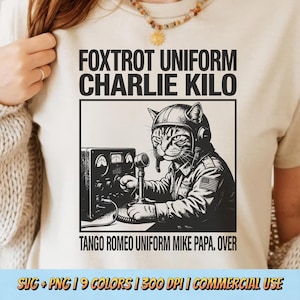 Op de afbeelding: Crèmekleurig T-shirt met een zwart-wit illustratie van een kat in militair uniform die een radio bedient. De tekst "FOXTROT UNIFORM CHARLIE KILO" en "TANGO ROMEO UNIFORM MIKE PAPA. OVER" is boven en onder de afbeelding gedrukt.