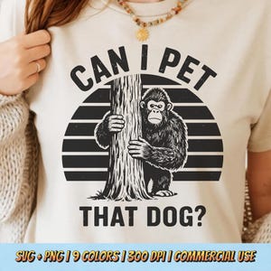 以下が含まれることがあります： ベージュ色のTシャツ。黒と白のゴリラが木の幹を抱きしめているグラフィックが、ストライプの背景に描かれています。「CAN I PET THAT DOG?」のテキストが画像の上と下に表示されています。