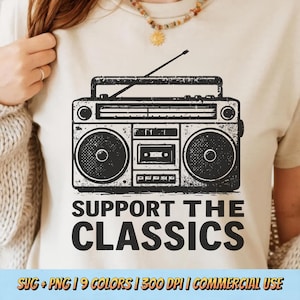 May include: A beige t-shirt featuring a black graphic of a vintage boombox with the text "SUPPORT THE CLASSICS". The design includes speakers, a cassette player, and an antenna. The shirt is a casual wear item.