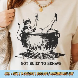 Könnte beinhalten: Beiges T-Shirt mit einer schwarz-weißen Illustration einer Teufelin, die sich in einem Kessel entspannt und ein Weinglas hält. Darunter steht der Text "NOT BUILT TO BEHAVE". Am unteren Rand des Shirts steht "SVG + PNG | 9 COLORS | 300 DPI | COMMERCIAL USE".