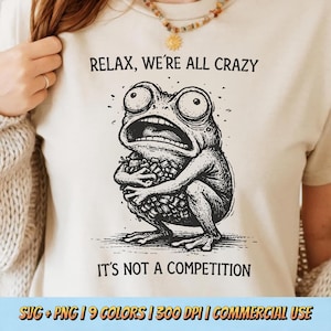 以下が含まれることがあります： ベージュ色のTシャツ。大きな目と開いた口をしたカエルのイラストが描かれています。「RELAX, WE'RE ALL CRAZY」の文字がカエルの上に、「IT'S NOT A COMPETITION」の文字が下にあります。Tシャツはニュートラルカラーです。