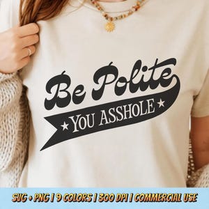 May include: Beige t-shirt with the phrase "Be Polite YOU ASSHOLE" in black script. The text is on a black banner with stars. The shirt is a casual style.