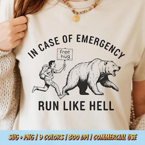 May include: Beige t-shirt with a black graphic of a person running from a bear. The text reads "IN CASE OF EMERGENCY" above the image and "RUN LIKE HELL" below. The person holds a sign that says "free hug".