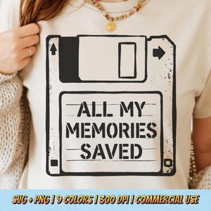 Op de afbeelding: Beige t-shirt met een zwart-witte afbeelding van een diskette en de tekst "ALL MY MEMORIES SAVED". Het ontwerp is in retrostijl. Het t-shirt heeft een ronde hals en korte mouwen.