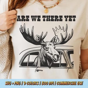 Op de afbeelding: Beige T-shirt met de tekst "ARE WE THERE YET" boven een zwart-wit illustratie van een eland die uit een autoraam kijkt. Het shirt heeft een ronde hals en korte mouwen.