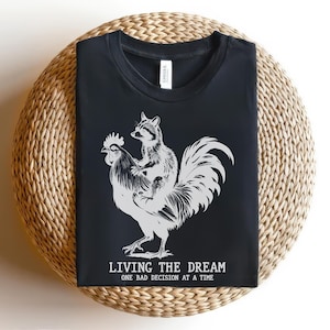 May include: Black t-shirt with a graphic of a raccoon riding a rooster. The text "LIVING THE DREAM ONE BAD DECISION AT A TIME" is printed below the graphic. The shirt is displayed on a woven, circular surface.