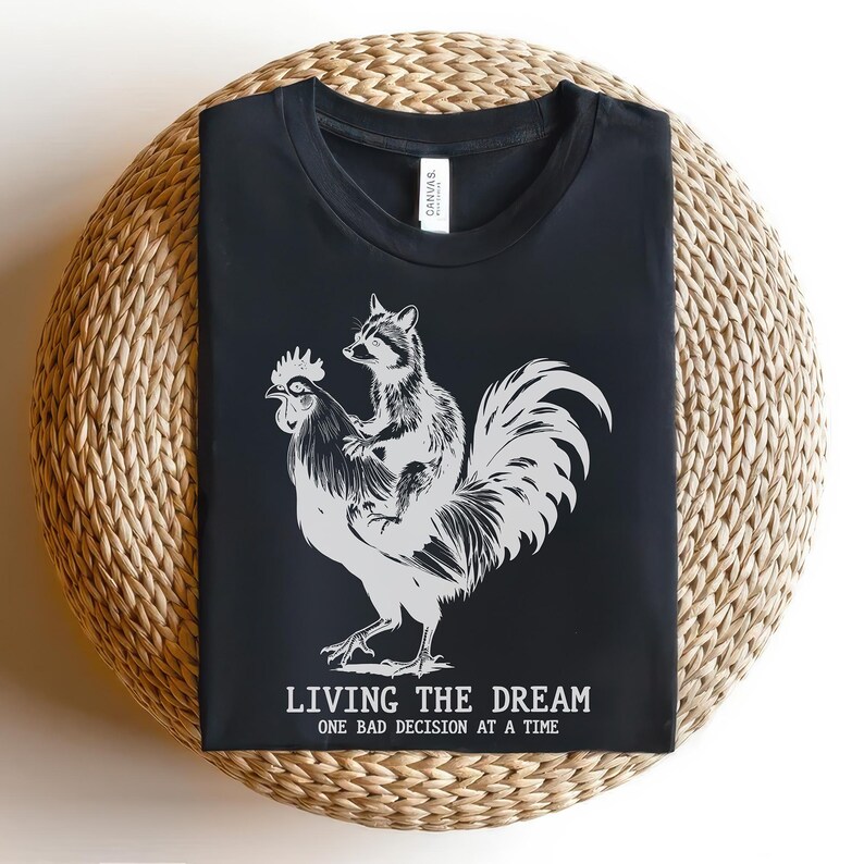 May include: Black t-shirt with a graphic of a raccoon riding a rooster. The text "LIVING THE DREAM ONE BAD DECISION AT A TIME" is printed below the graphic. The shirt is displayed on a woven, circular surface.