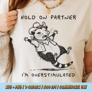 Könnte beinhalten: Beiges T-Shirt mit einer schwarz-weißen Illustration eines Opossums mit Cowboyhut und Weste, mit dem Text "HOLD ON PARTNER I'M OVERSTIMULATED". Das Design ist humorvoll.
