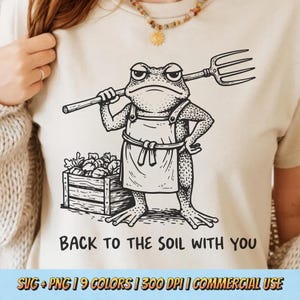May include: Beige t-shirt with a black and white illustration of a frog holding a pitchfork and wearing an apron. The text "BACK TO THE SOIL WITH YOU" is below the frog. The shirt has a crew neck.