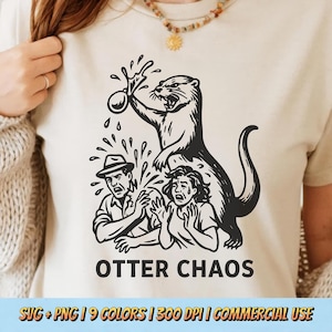 May include: Beige t-shirt with a black graphic of an otter holding a water balloon above two people. The text "OTTER CHAOS" is below the graphic. The shirt is a light neutral color.