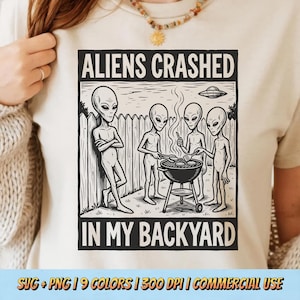 Puede incluir: Camiseta color crema con un gráfico en blanco y negro de extraterrestres haciendo una barbacoa. El texto dice "ALIENS CRASHED IN MY BACKYARD". El diseño incluye una valla y un OVNI en el fondo.