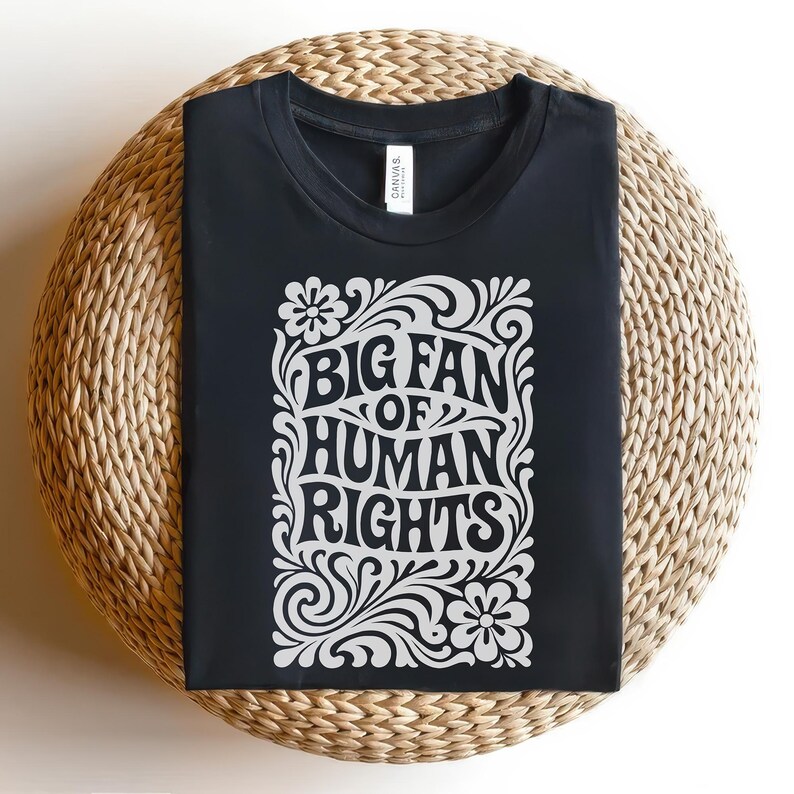 Puede incluir: Camiseta negra con el texto blanco "BIG FAN OF HUMAN RIGHTS" en un dise&ntilde;o floral. La camiseta se muestra sobre una superficie redonda y tejida.
