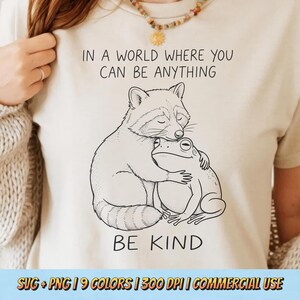 May include: Cream-coloured t-shirt featuring a black and white drawing of a raccoon embracing a frog. The words "IN A WORLD WHERE YOU CAN BE ANYTHING BE KIND" are printed above and below the image. The bottom of the shirt has the text "SVG + PNG | 9 COLORS | 300 DPI | COMMERCIAL USE".