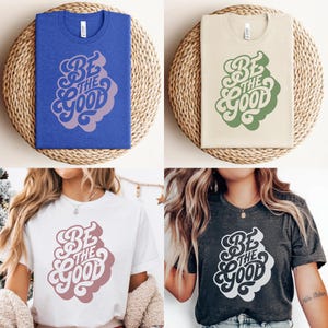 May include: Four t-shirts in different colors: blue, beige, white, and charcoal gray. Each shirt features the text "Be the Good" in a retro-style design. The shirts are displayed on woven circular mats.